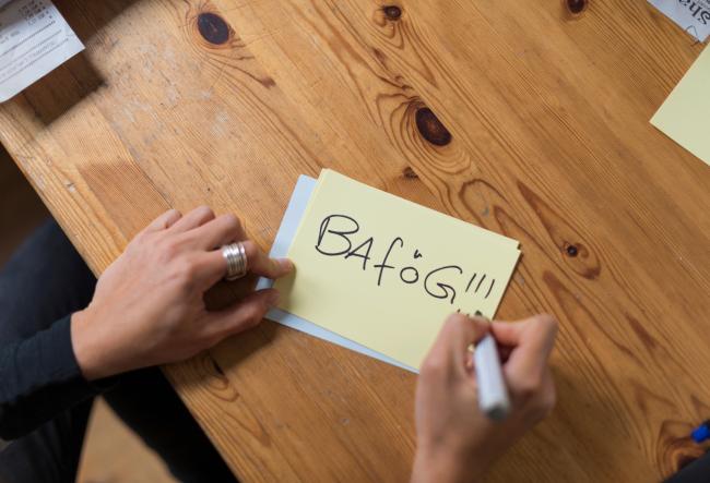 A hand can be seen writing ‘Bafög!!!’ on a yellow piece of paper with a black marker.