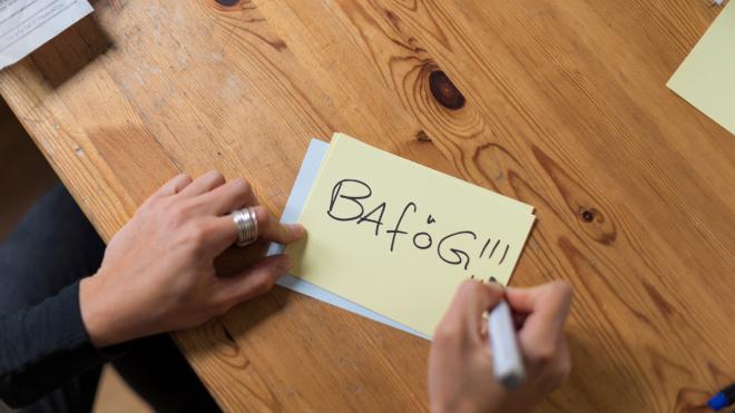 A hand can be seen writing ‘Bafög!!!’ on a yellow piece of paper with a black marker.