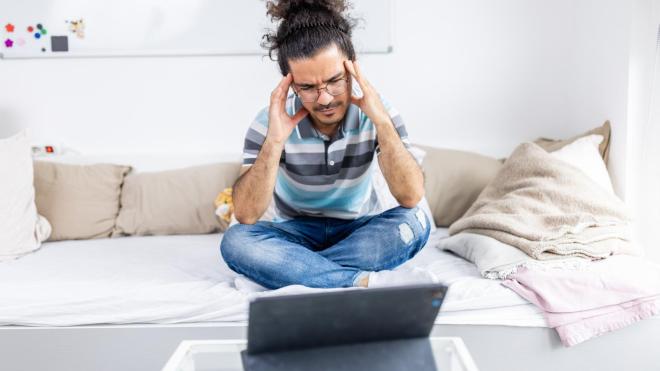 A stressed man sits on the bed and holds his head. 