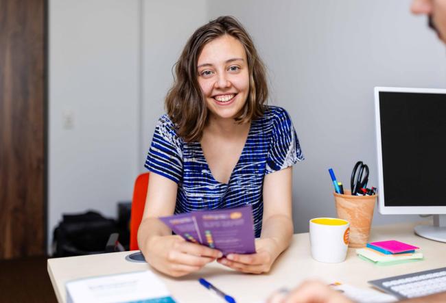 Clerk in her office with a student, holding a flyer and smiling at the camera.