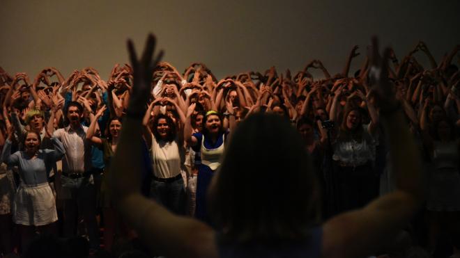 A Choir is singing and forming hearts with their hands above their head. The conductor is standing large in the foreground and raising her hands.