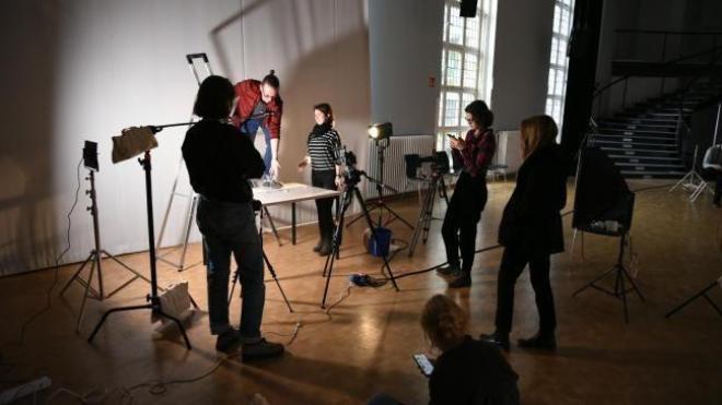 Six people stand in a circle in the open space surrounded by lighting technology, one of them stands on a ladder and points down to a table.