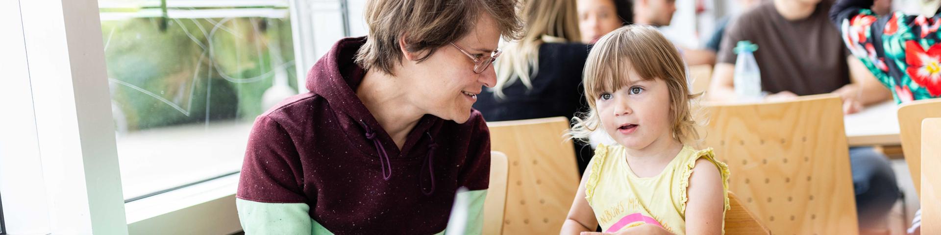 Student smiles at her toddler. They sit together at a table in a canteen and eat.