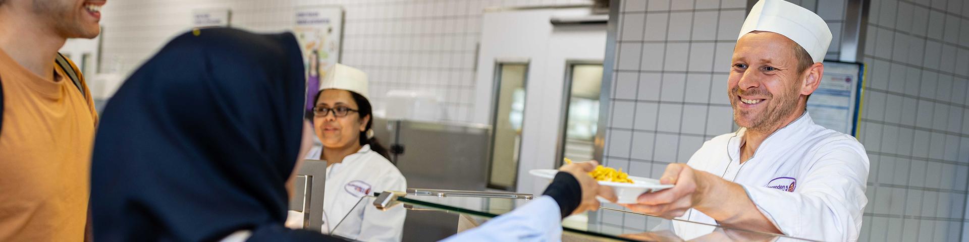 Mensa employee hands a meal over the counter with a smile.