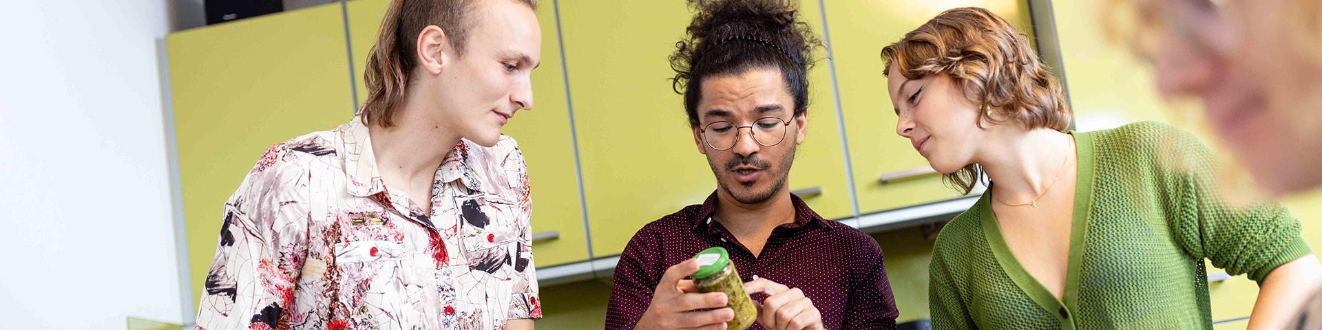 Students cooking in a hall of residence kitchen. Together they look at the ingredients in a jar of food.