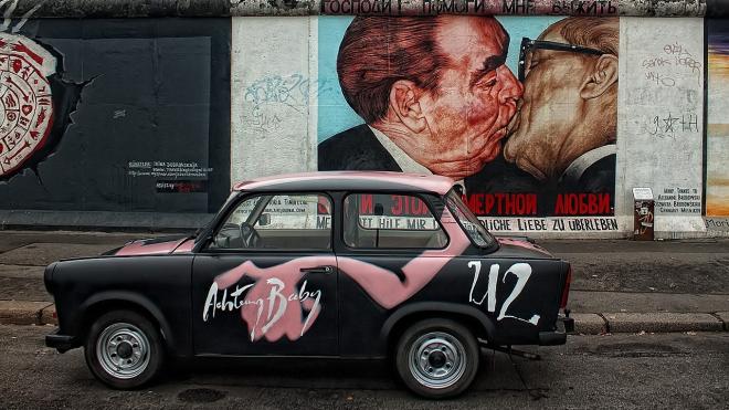 Mauerstück der Berliner Mauer zeigt den ikonischen „Bruderkuss“ zwischen Breschnew und Honecker, mit einem Trabant davor