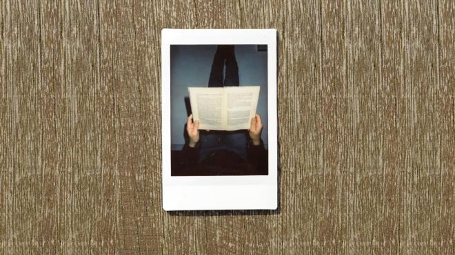 Ein Polaroid von einer Person, die entspannt ein Buch auf den Knien hält, liegt auf einem Tisch. 