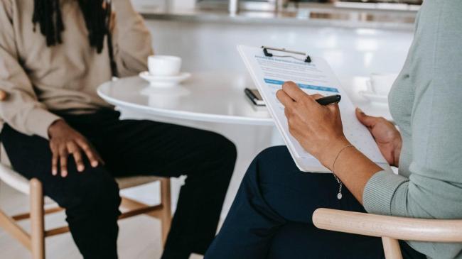 Photo shows two people at a table with coffee in an interview situation, one person is taking notes.