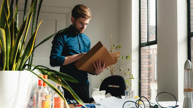 A man is standing in front of a messy desk holding a folder in his hands.